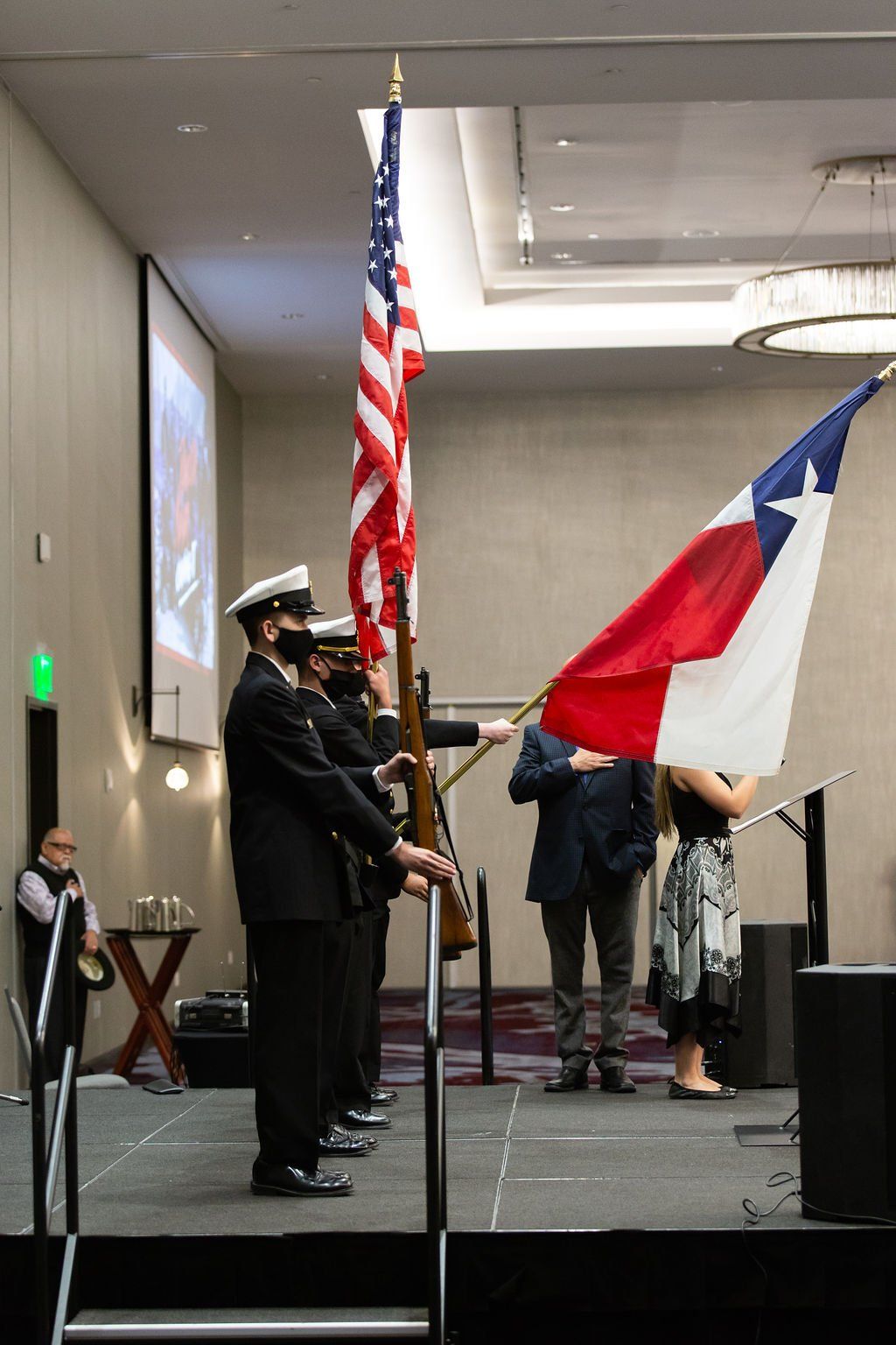 A color guard stands on stage with the U.S. and Texas flags, with individuals behind them observing in a formal setting.