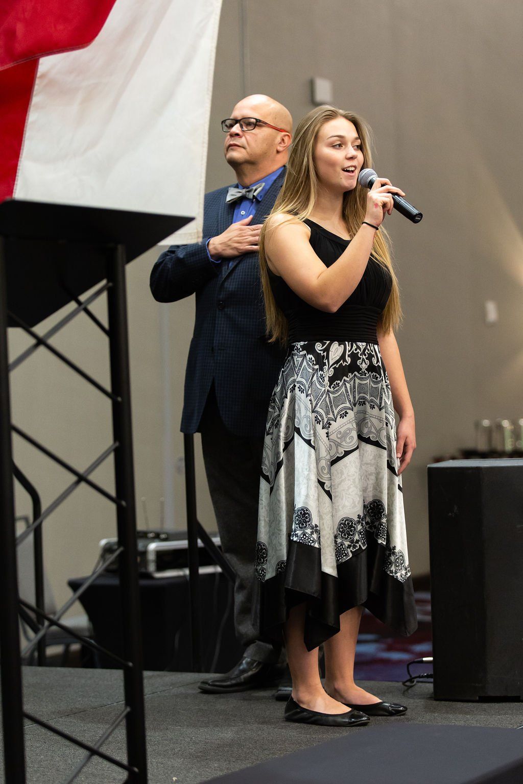 A woman speaks into a microphone on stage while a man stands behind her with his hand over his heart near a flag.