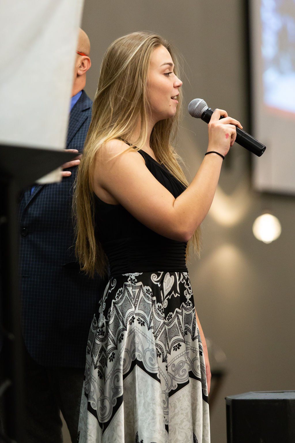 A person with long blonde hair speaks into a microphone while wearing a black top and a black-and-white patterned skirt.