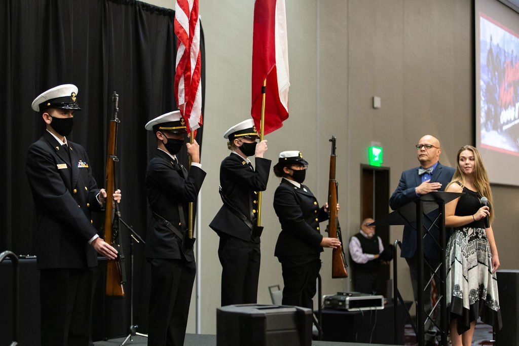 A color guard stands on stage with flags and rifles, flanked by two people, one with a hand over their heart.