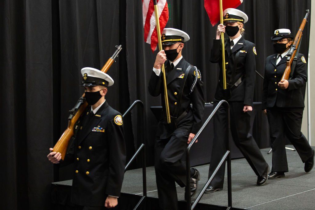 Color guard members in dress uniforms and face masks march with flags and rifles on a stage with a black backdrop.