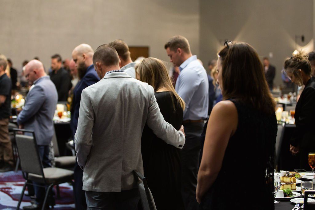 A crowd stands with heads bowed in a dimly lit banquet hall, many people with their arms around each other.