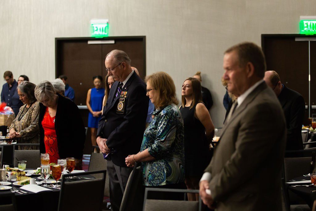 A group of people standing with heads bowed in a silent moment at a formal event in a banquet hall.