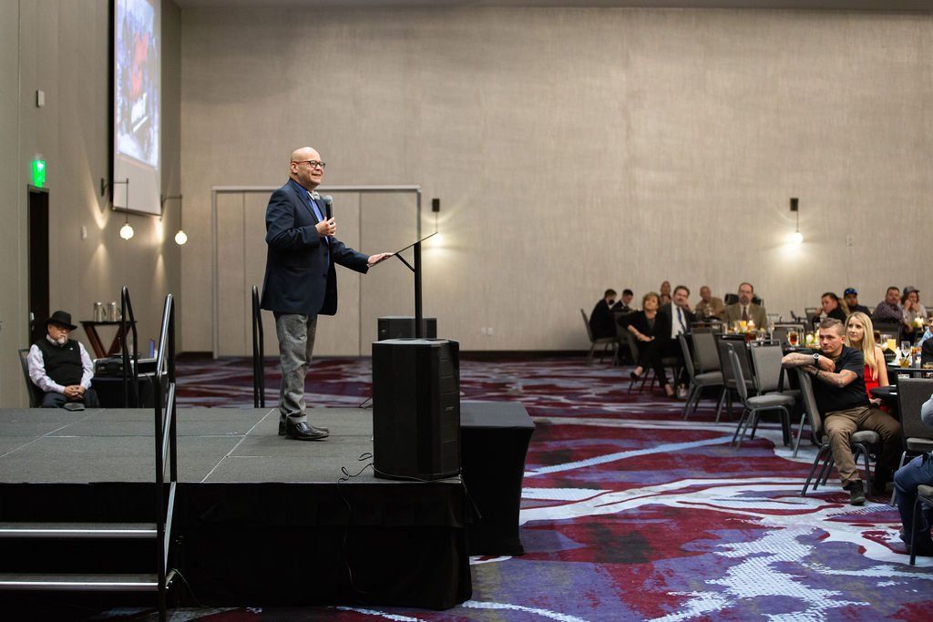 A person standing on a stage at a podium addressing an audience seated at tables in a large event hall.