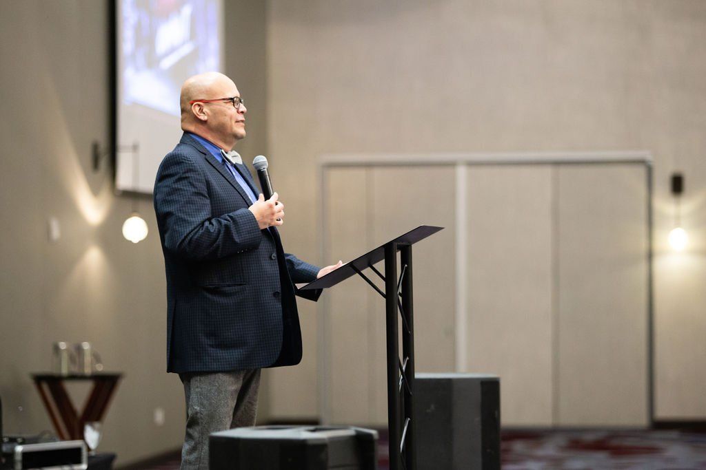 A speaker in a dark blazer stands at a lectern on stage holding a microphone, addressing an audience in a hall.