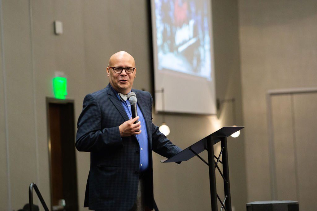 A person in a blue shirt and dark blazer holds a microphone while speaking from a lectern in a room with a projection.