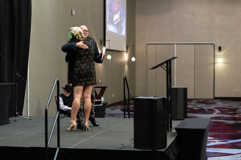Two people in formal attire embrace on a stage during an indoor event.