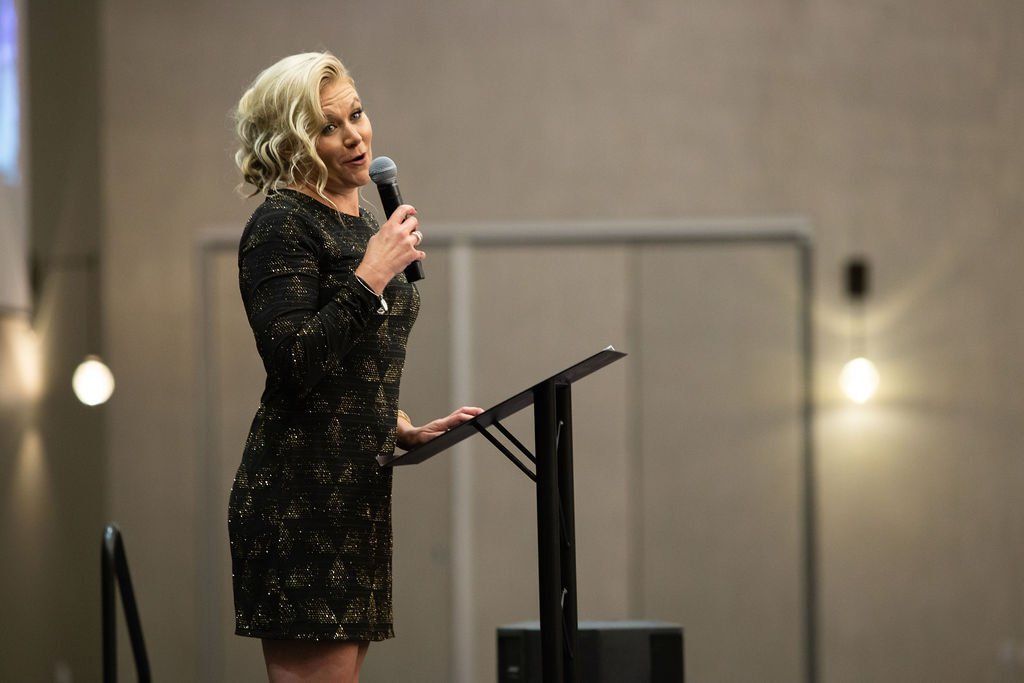A blonde woman speaking into a microphone from a podium in a modern, softly lit hall.