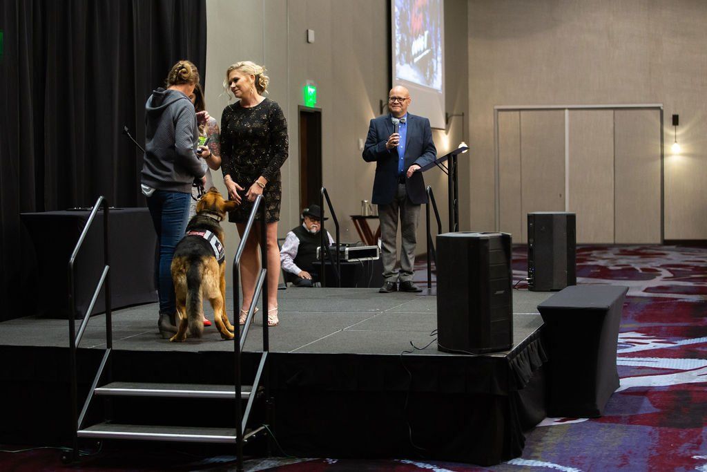 People stand on a stage with a German Shepherd; a person in a suit speaks into a microphone at an event.