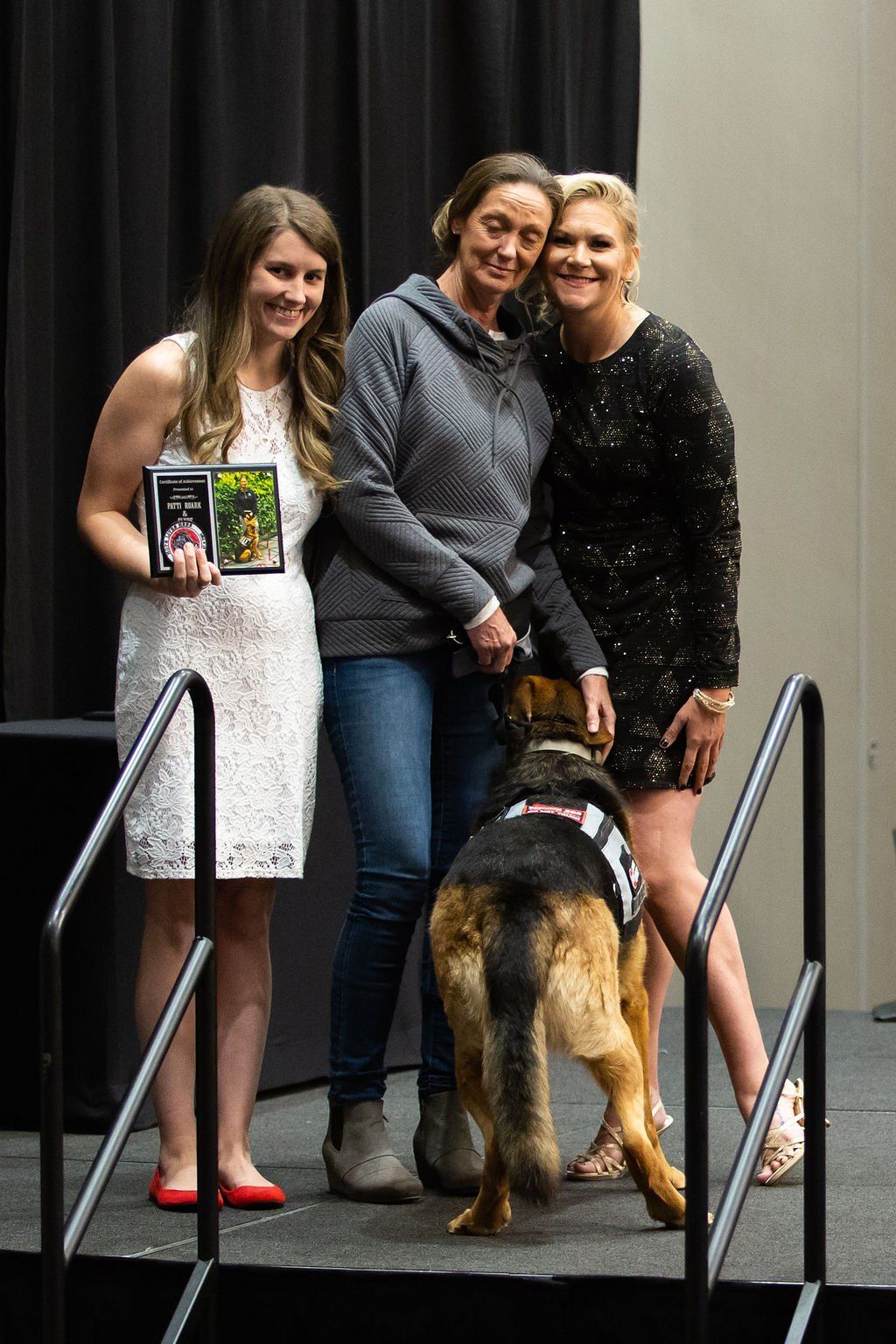 Three people stand on a stage with a German Shepherd; one person holds a plaque, while the dog faces away from the camera.