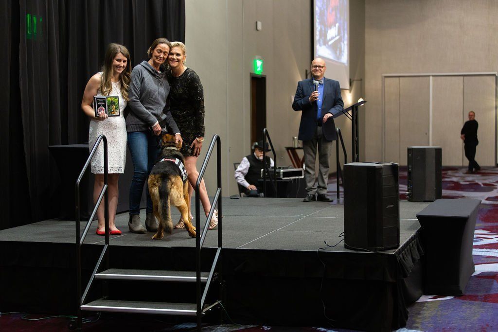 Three people and a German Shepherd stand on a stage during an award presentation, with a speaker at a podium nearby.