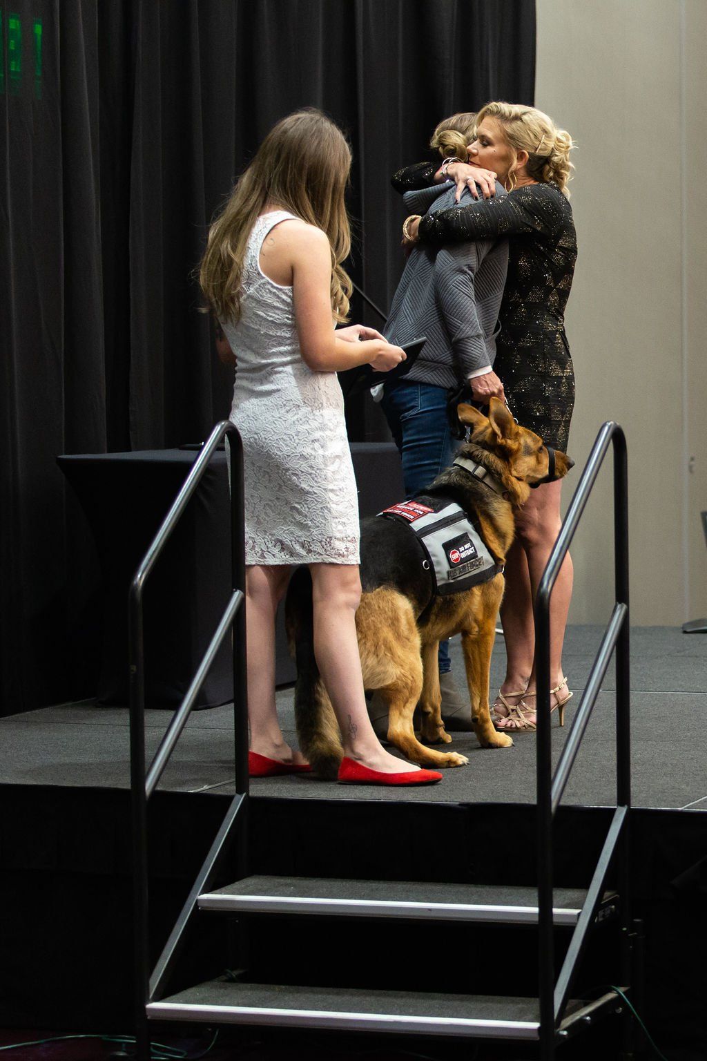 A service dog with a harness stands on a stage next to three people, two of whom are embracing while one stands nearby.