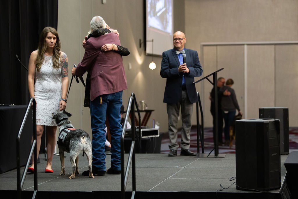On a stage, two people embrace while a woman and a service dog stand nearby, with a man standing at a podium to the right.