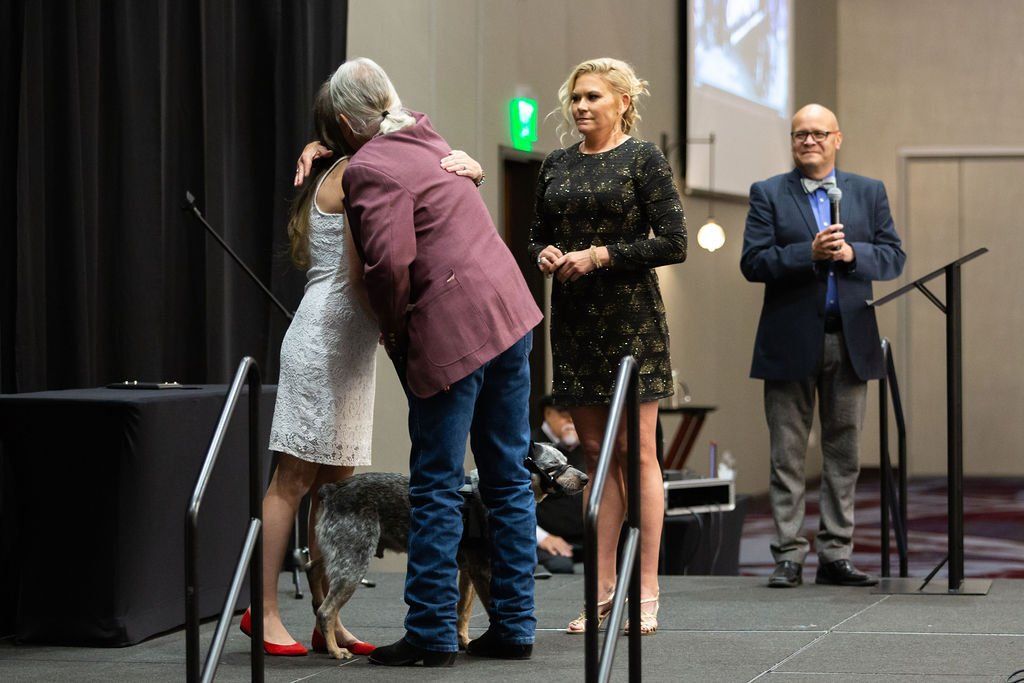 A person in a maroon jacket hugs someone in a white dress on a stage, next to a woman in a black dress and a man in a suit.