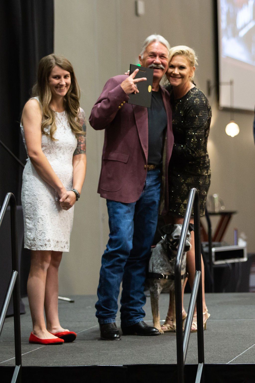 Three people and a dog stand on a stage. A person in a maroon blazer holds a plaque while smiling with two others.