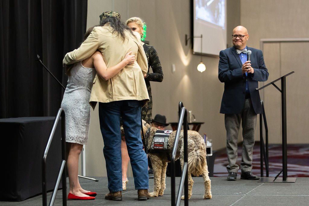 Two people hug on a stage next to a service dog while a person in a suit stands nearby and claps.