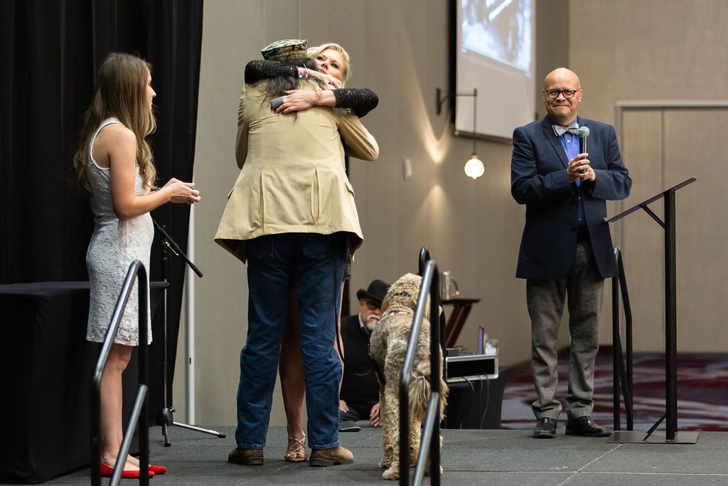 A person in a beige jacket and blue jeans hugs another person on a stage, watched by a person in a suit and a child.