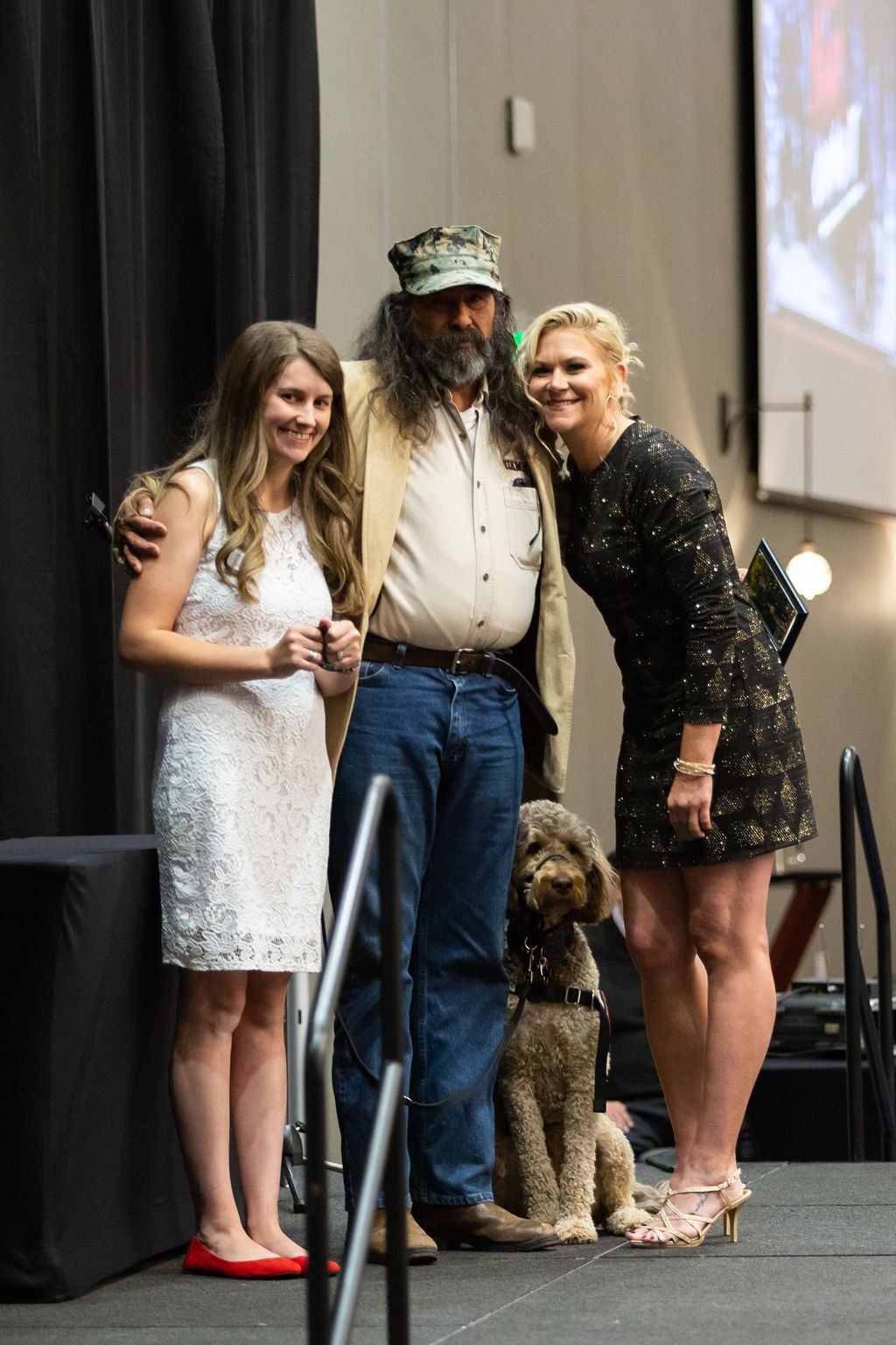 Three people and a dog stand together on an indoor stage, posing for a photo against a backdrop of black curtains.