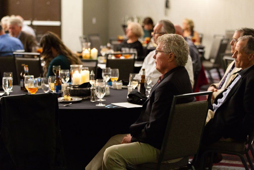People seated at round tables in a conference room, listening attentively to a presentation.