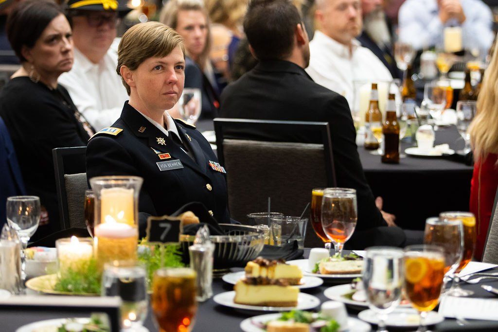 A person in a military dress uniform sits at a formal dinner table, looking forward among other guests in a dimly lit room.
