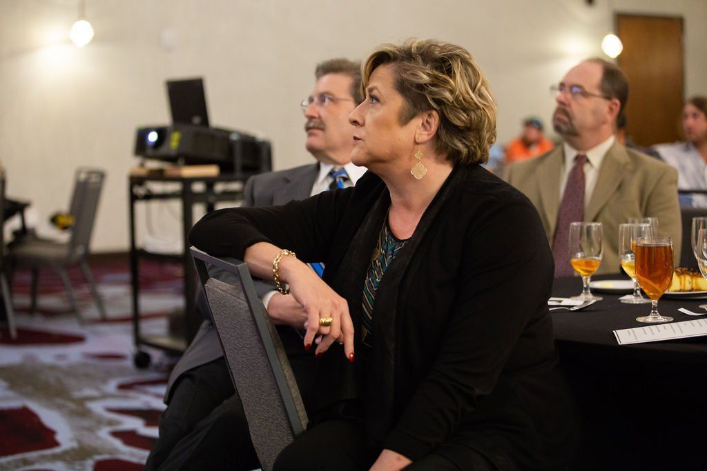 People in formal attire listen attentively during a presentation in a conference room with a projector in the background.