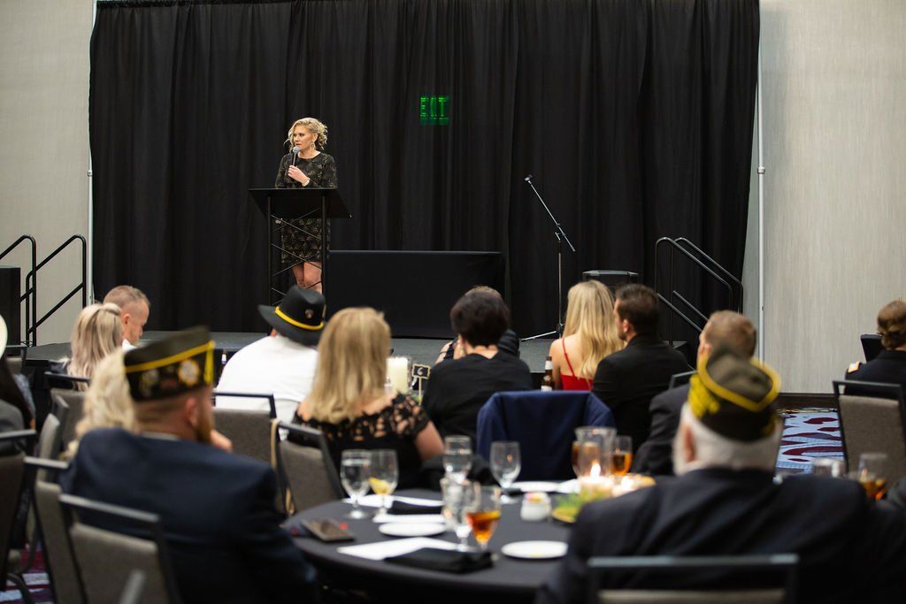 A woman speaks at a podium on a stage with black curtains, addressing an audience seated at round tables in a formal hall.