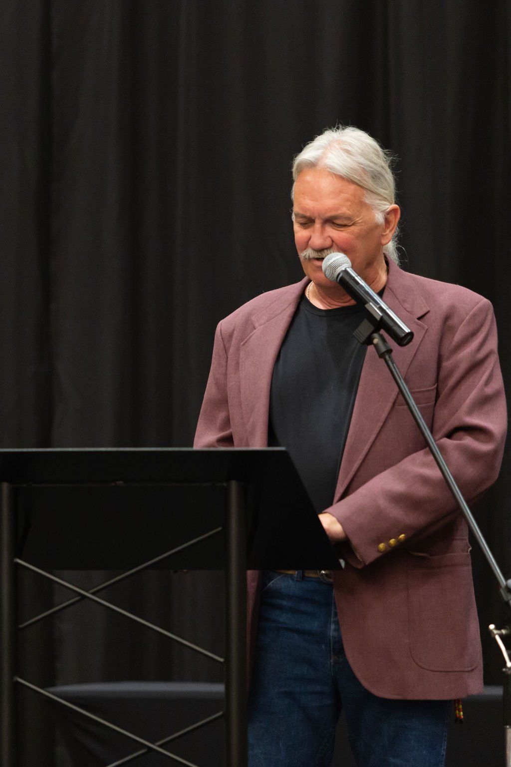 A person in a maroon blazer speaks into a microphone at a podium in front of a black curtain.