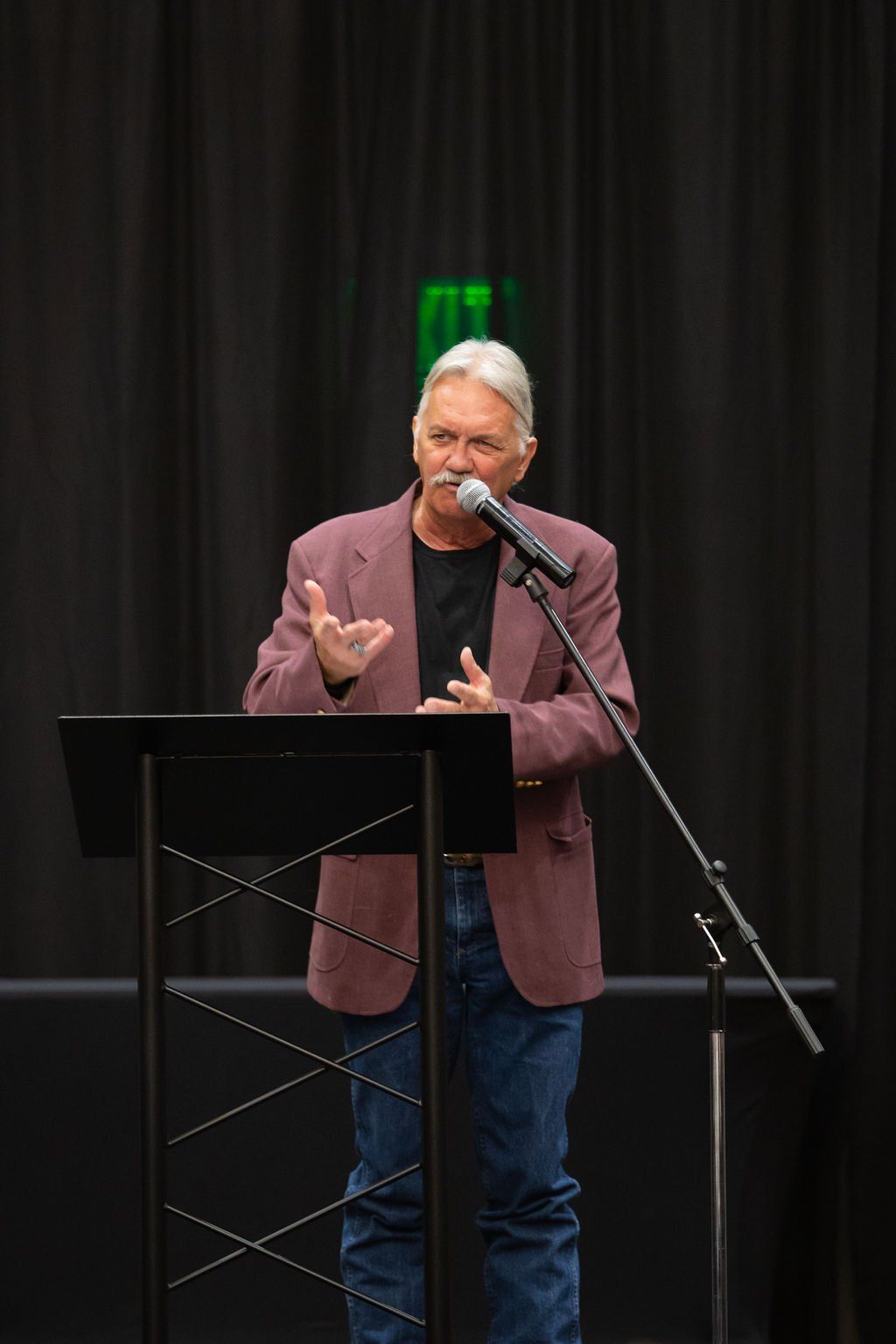 A person in a maroon blazer speaks at a lectern with a microphone in front of a dark curtain.