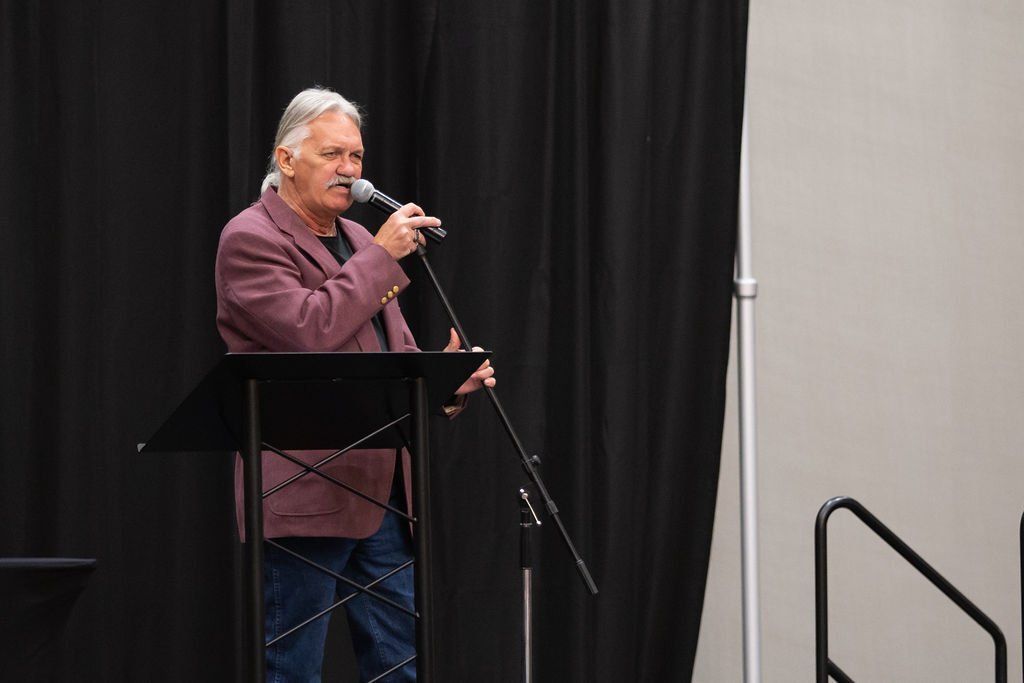 A person in a maroon blazer stands at a podium, speaking into a microphone in front of black curtains.