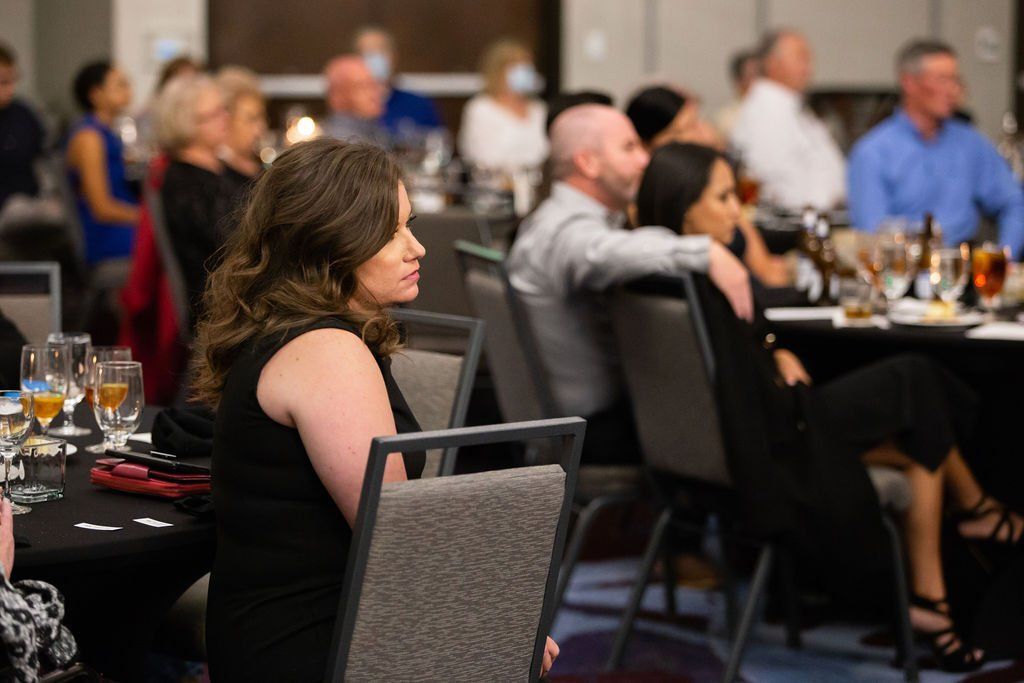 People seated at tables in a dimly lit conference room, listening to a speaker or presentation.