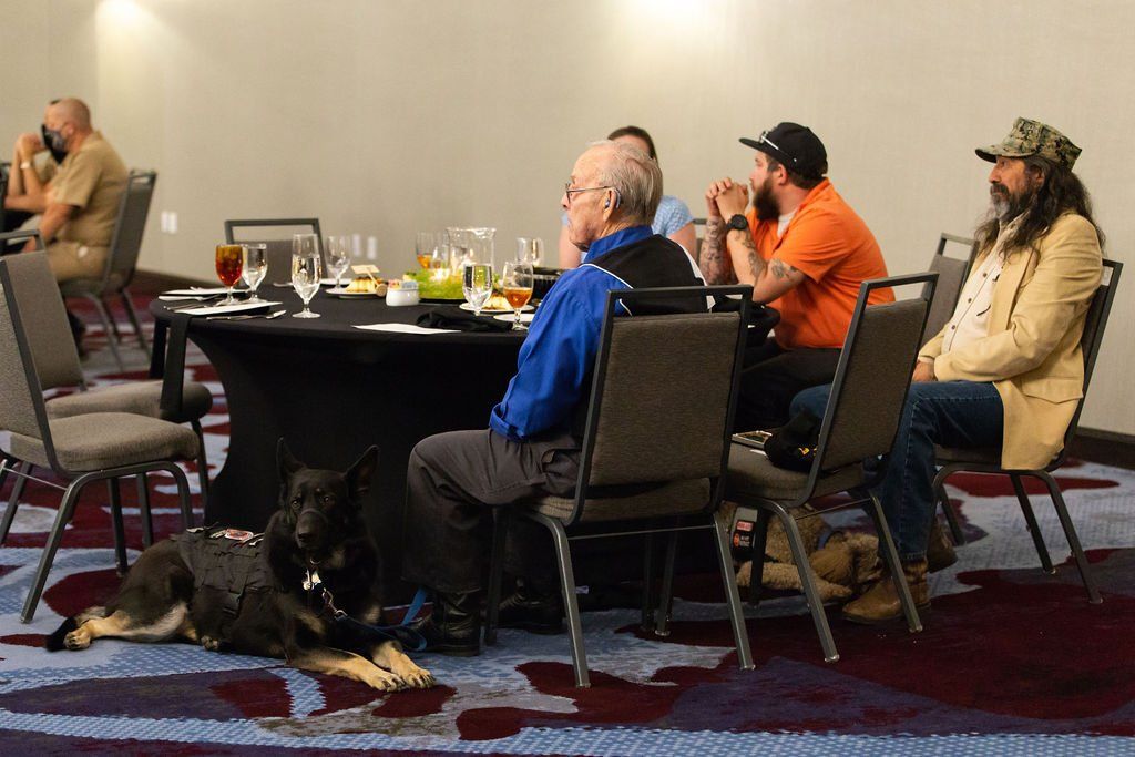 A service dog lies on the floor under a dining table as four people sit and attend a meeting in a conference room.