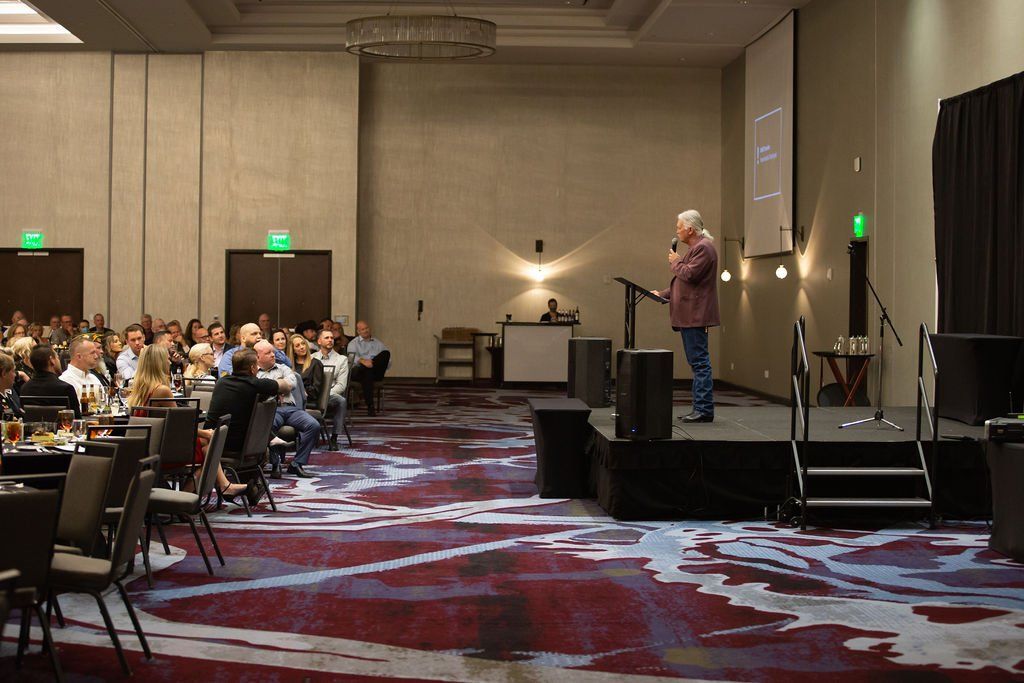 A speaker stands on a stage at a podium addressing an audience seated at tables in a conference room with patterned carpet.