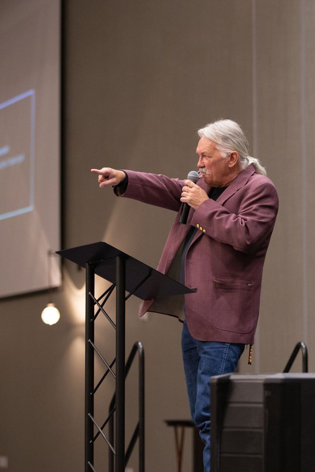 A person in a maroon blazer stands at a podium, speaking into a microphone and gesturing to the side in a large room.