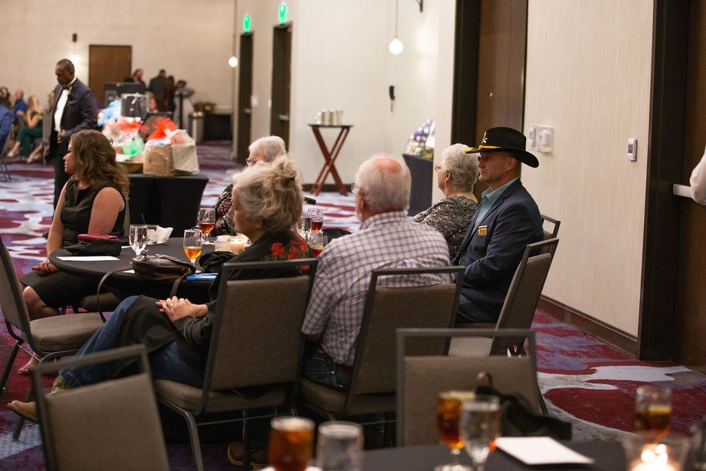 People sit at tables in a brightly lit banquet hall, facing the front of the room during an event.