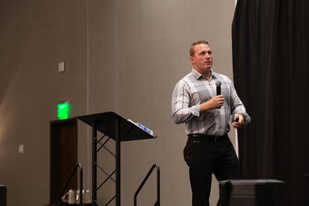 A speaker in a button-down shirt holds a microphone while presenting on a stage next to a podium.