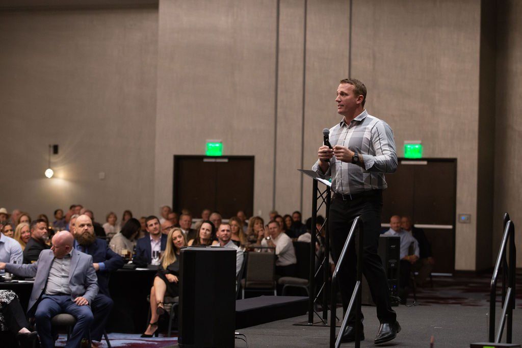 A speaker stands at a podium addressing an audience in a conference room with dimly lit exits.