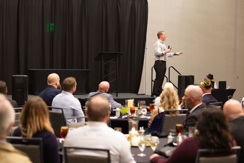A person stands on a stage presenting to an audience seated at round tables in a conference room.