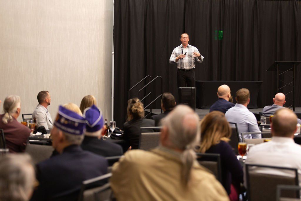 A speaker stands at a podium in a conference room, presenting to an audience seated in rows of chairs facing them.