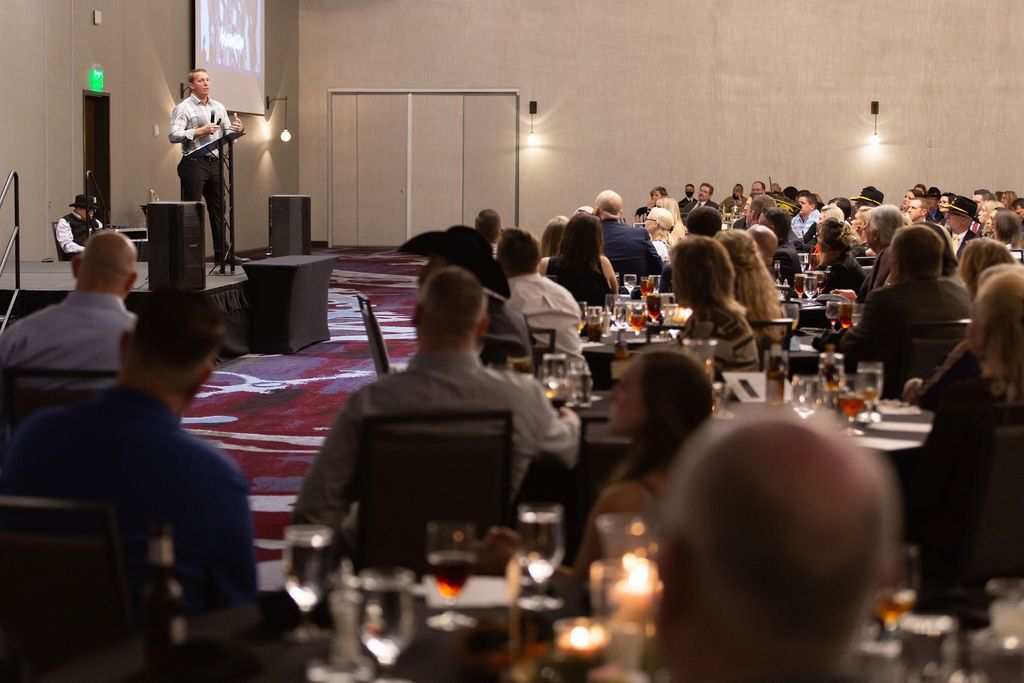 A speaker stands at a podium in a hotel ballroom addressing a seated audience during a banquet or formal event.
