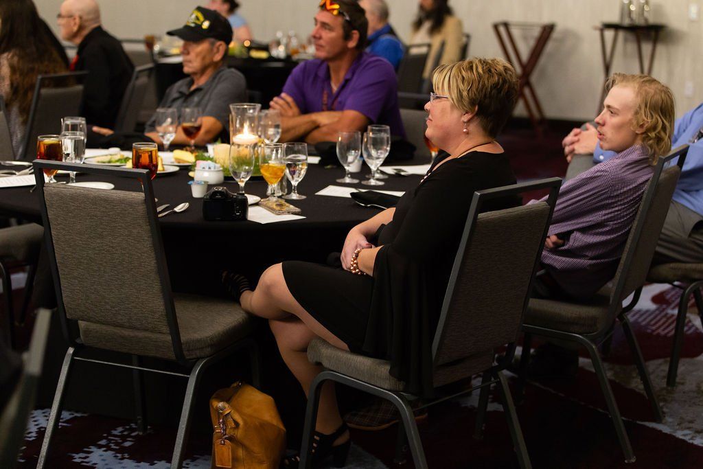 A group of people sit around a black-clothed table at a formal event, listening to a speaker.