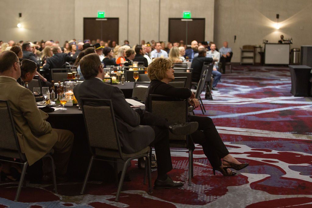 People sit at tables in a large event ballroom with a patterned carpet, facing a presentation.
