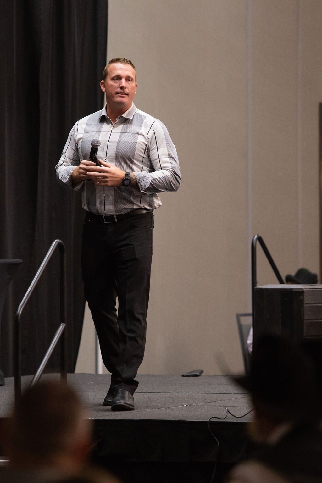 A speaker stands on a stage holding a microphone, wearing a light-colored button-down shirt and dark slacks.