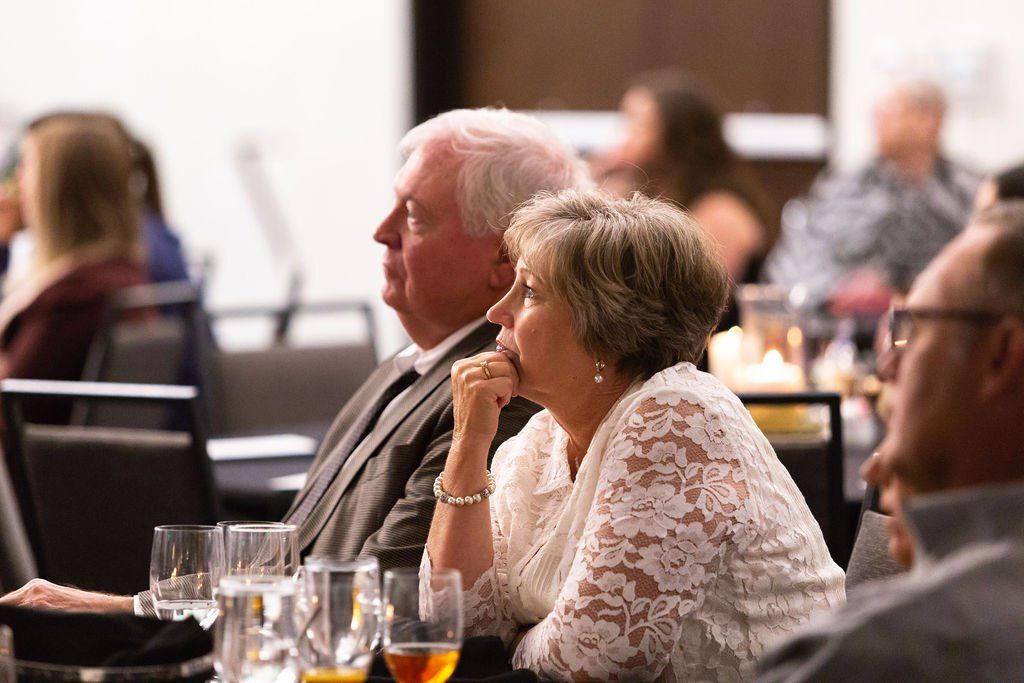 Two people sit at a table in a dimly lit room, attentively watching an event.