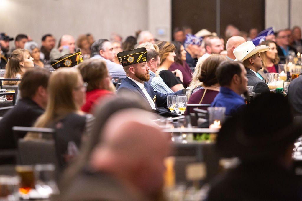 A large group of people sit at tables in a banquet hall, some wearing veterans' caps, listening to a presentation.