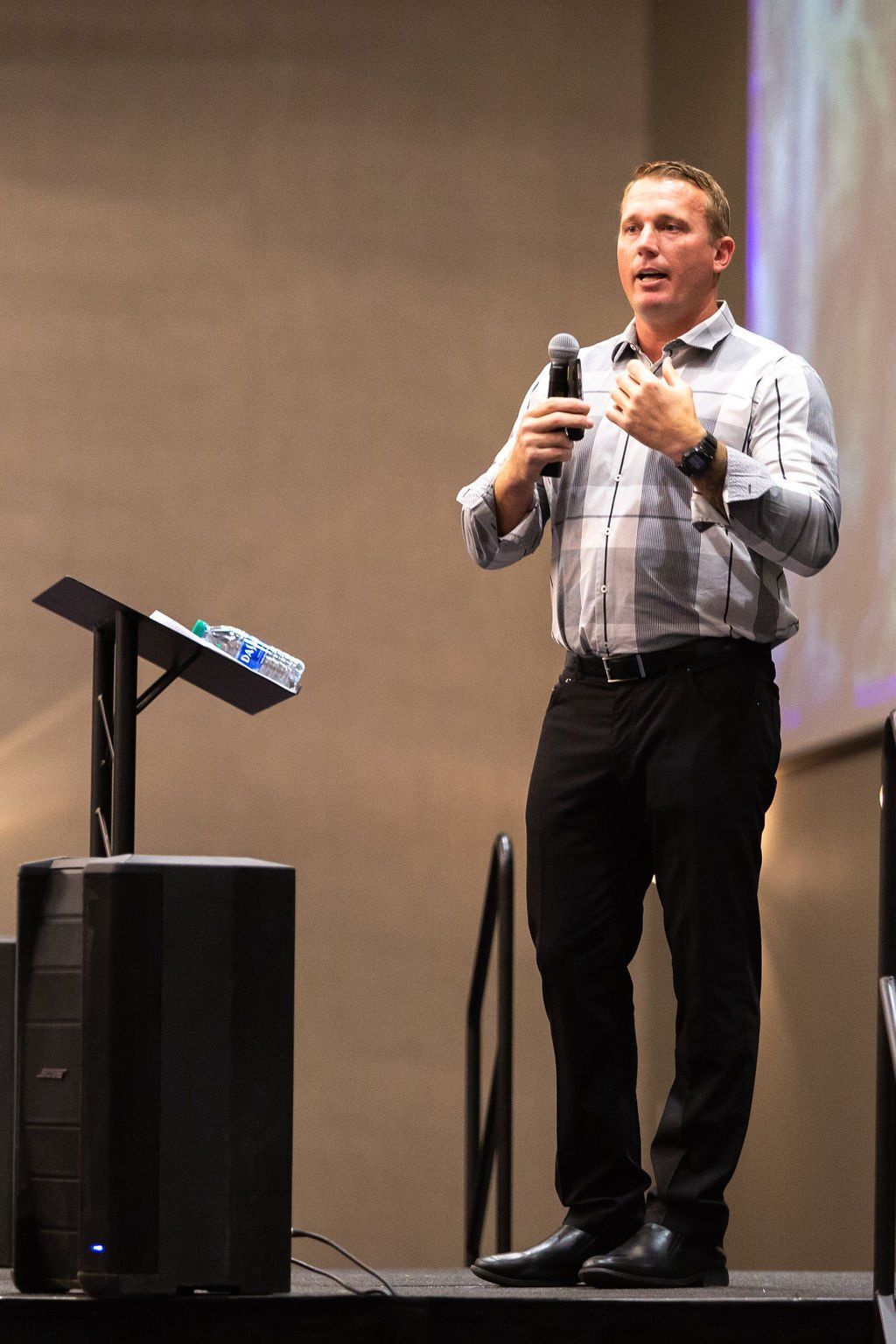 A speaker in a button-down shirt stands at a podium holding a microphone, gesturing while presenting on a stage.