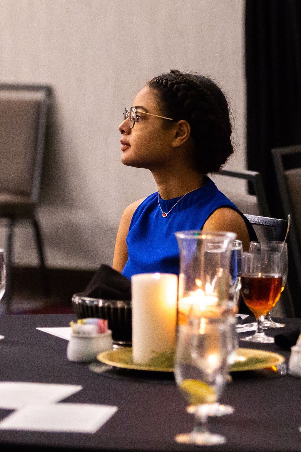 A person with braided hair and glasses in a blue sleeveless top looking to the side at a table with candles and glassware.