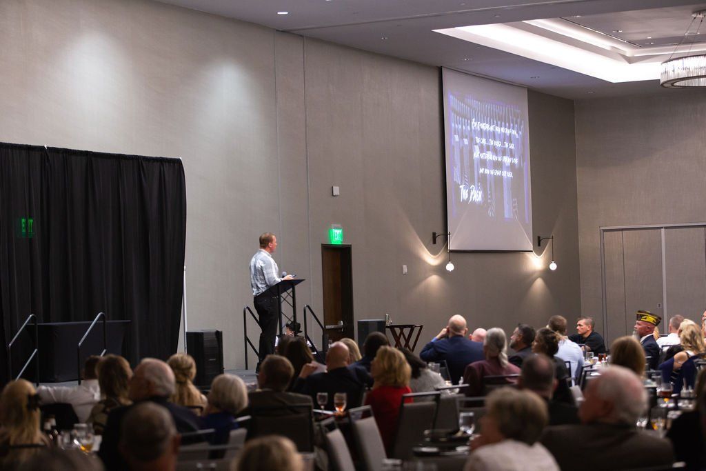 A speaker at a podium addresses a seated audience in a conference room with a presentation displayed on a projection screen.