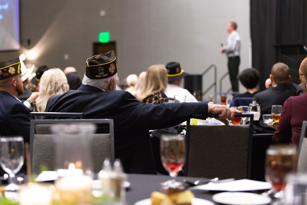 A banquet hall scene with attendees, some wearing veterans' caps, facing a speaker on a stage.