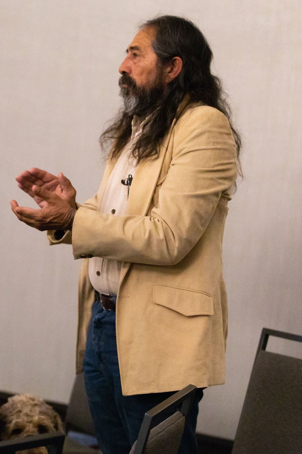 A long-haired person with a beard, wearing a tan blazer and jeans, claps while standing in a room with chairs.