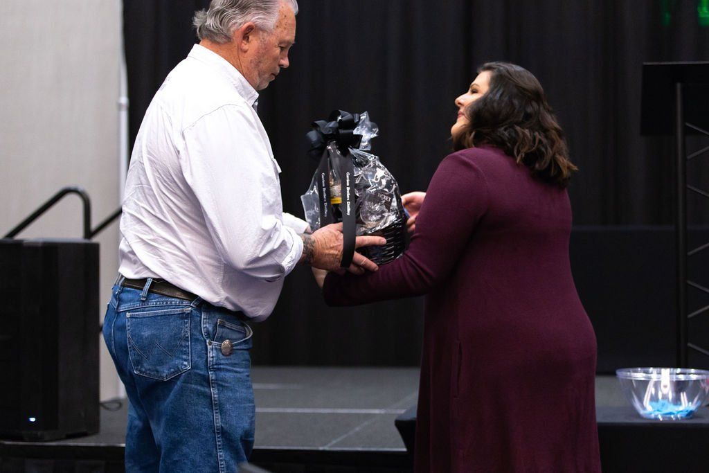 A man in a white shirt and jeans hands a gift wrapped in clear plastic to a person in a long-sleeved burgundy dress.